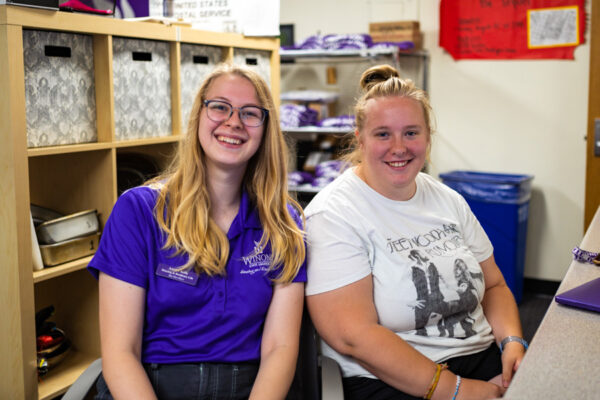 Two student workers smile while sitting behind the front desk in Sheehan Hall.