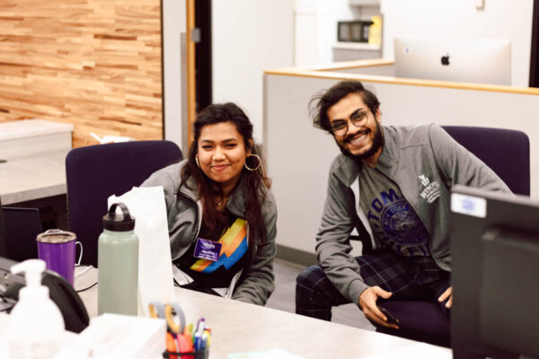 Two student workers smile while sitting behind a desk in an office at WSU.